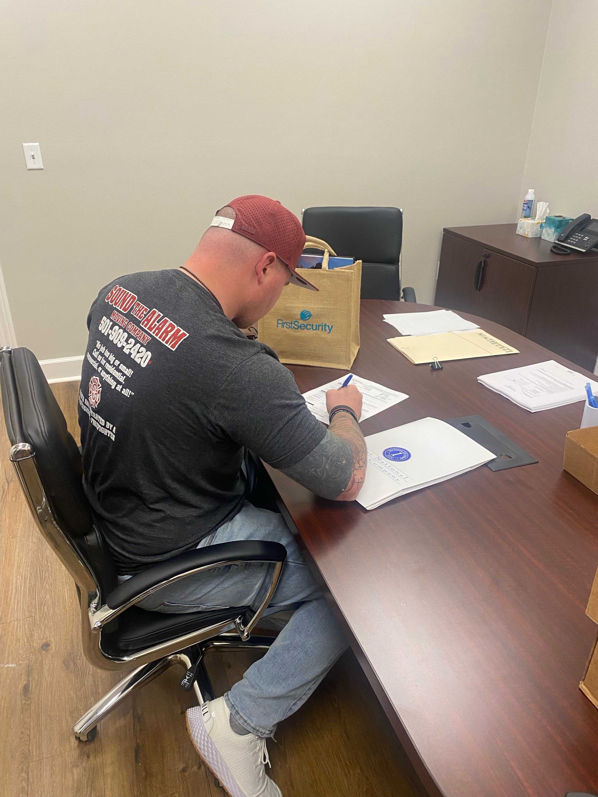 Person in a baseball cap seated at a desk, writing on paper. Office setting, brown desk, and black chair.