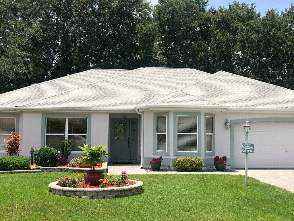 Single-story white house with a gray roof, front lawn, flower beds, and a driveway.