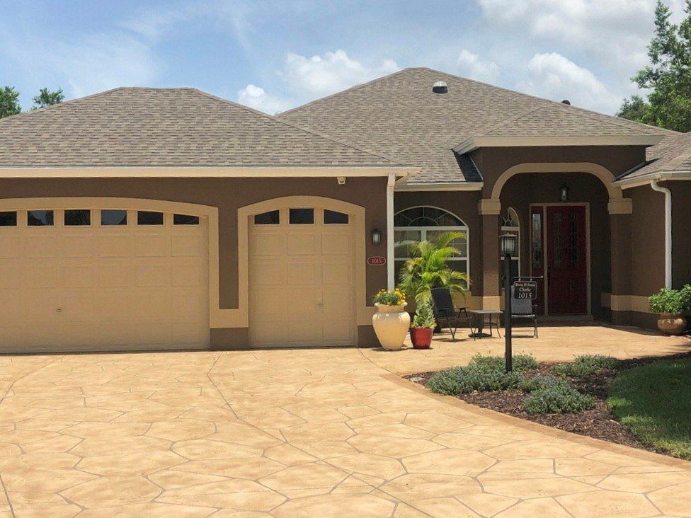 Suburban house with tan garage doors, stone driveway, and front porch under a partly cloudy sky