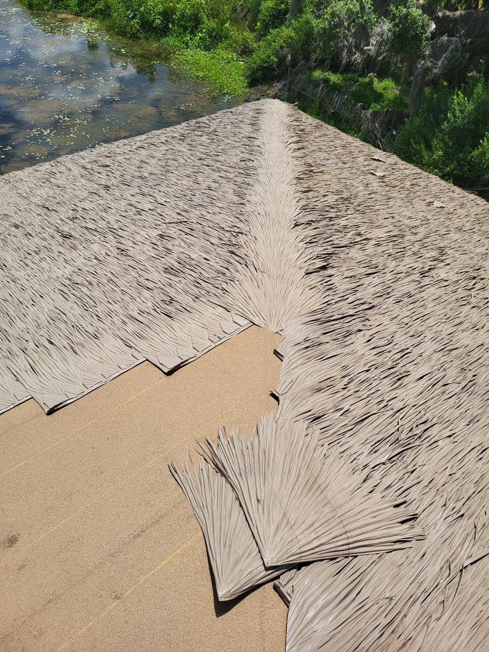 Thatched roof with a small repaired patch, viewed beside a body of water and green vegetation.