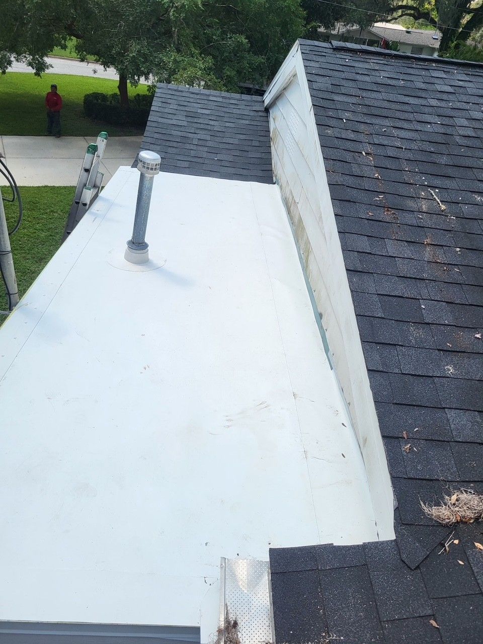 White flat roof beside dark shingle roof, with a metal vent pipe and a person on a lawn in the background