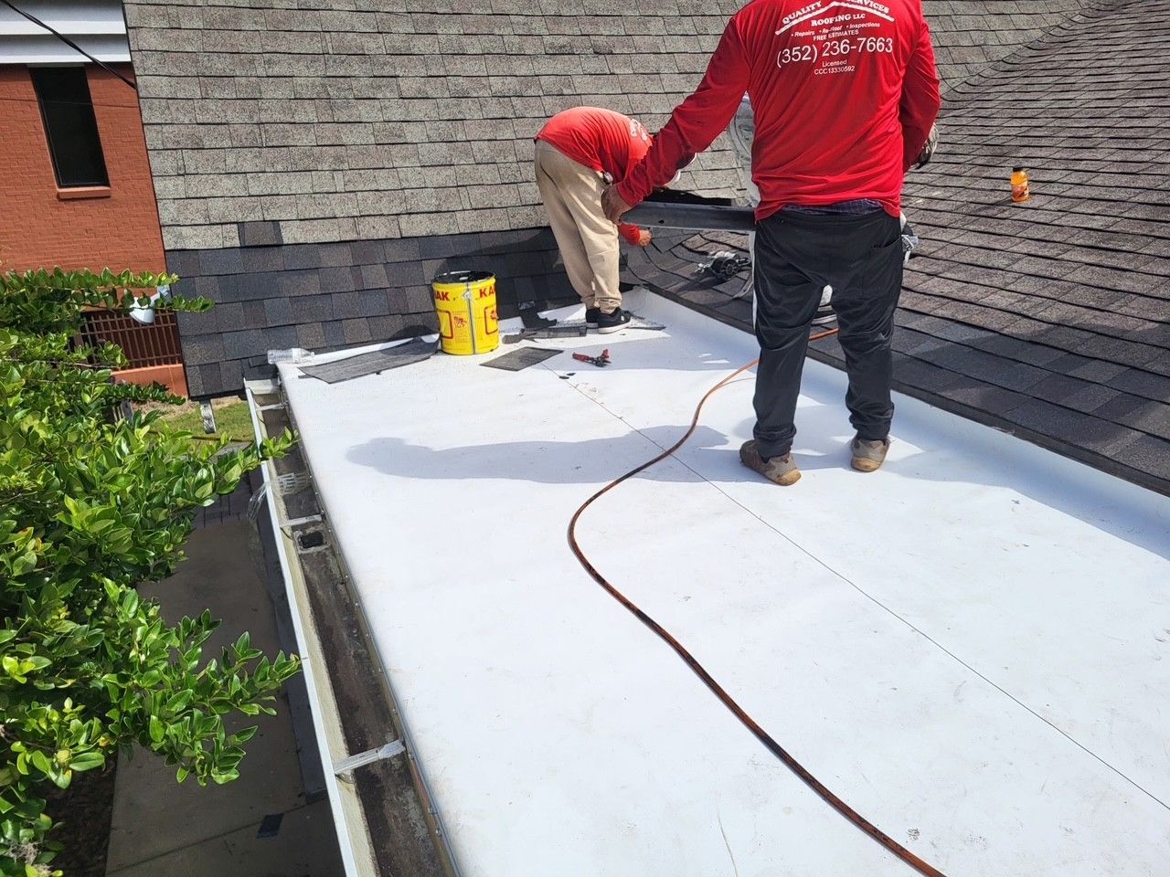 Two workers installing a white roof membrane on a rooftop, with tools and a yellow bucket nearby.