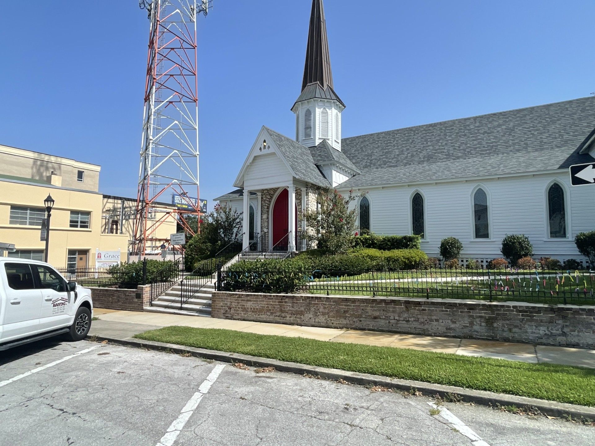 White church with tall steeple beside a parking lot, stone steps, and a red tower under a blue sky