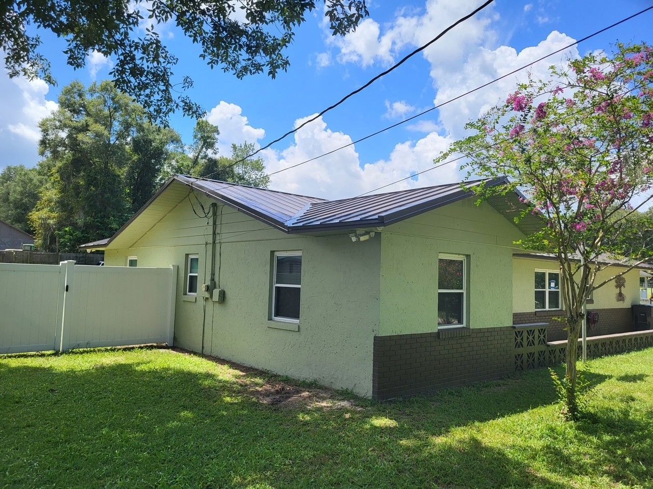 Single-story green house with a white fence and solar panels, viewed from a grassy yard under blue sky.