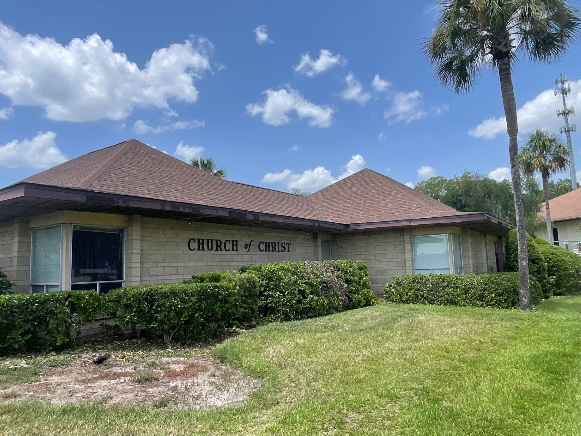 Church building with a brown roof, stone exterior, hedges, and palm trees under a blue sky.