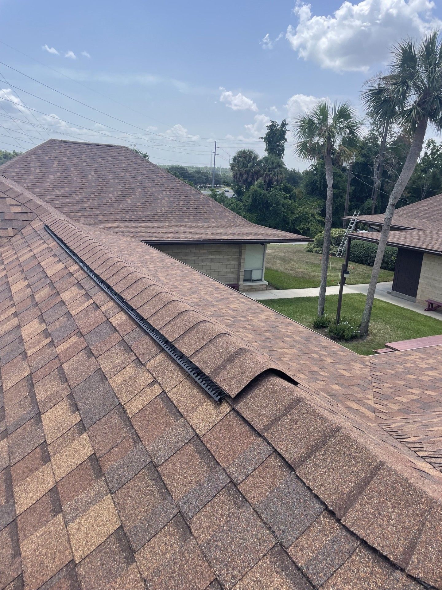 Rooftop with brown shingles and a ridge vent overlooking a green yard and cloudy sky