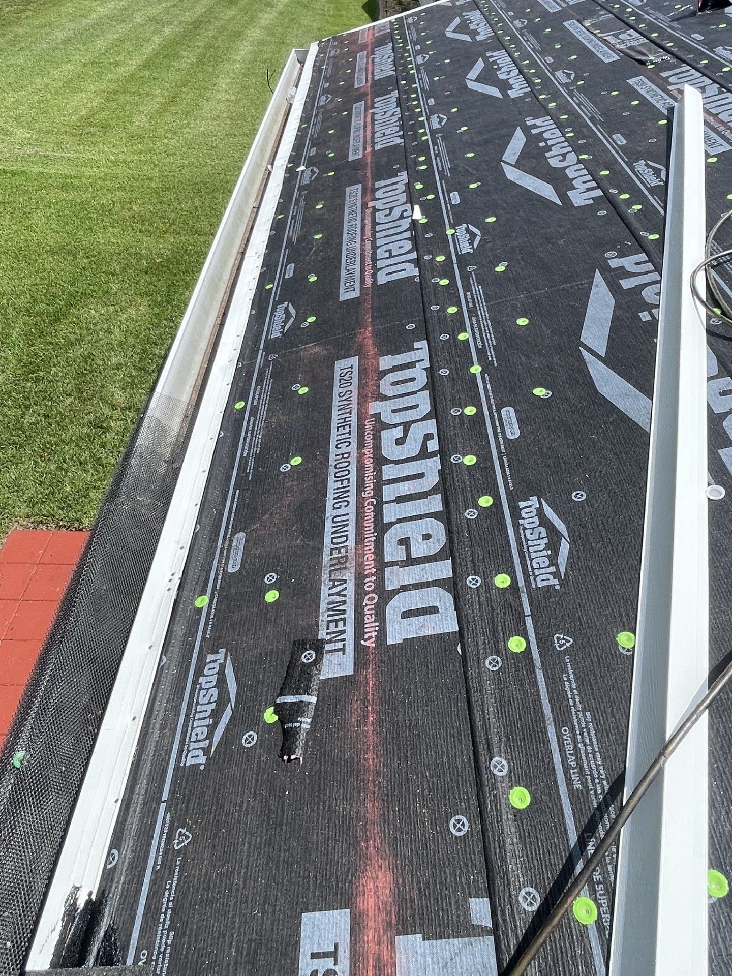 Roof with black DECKSHIELD underlayment, green fasteners, and a white railing beside grass.