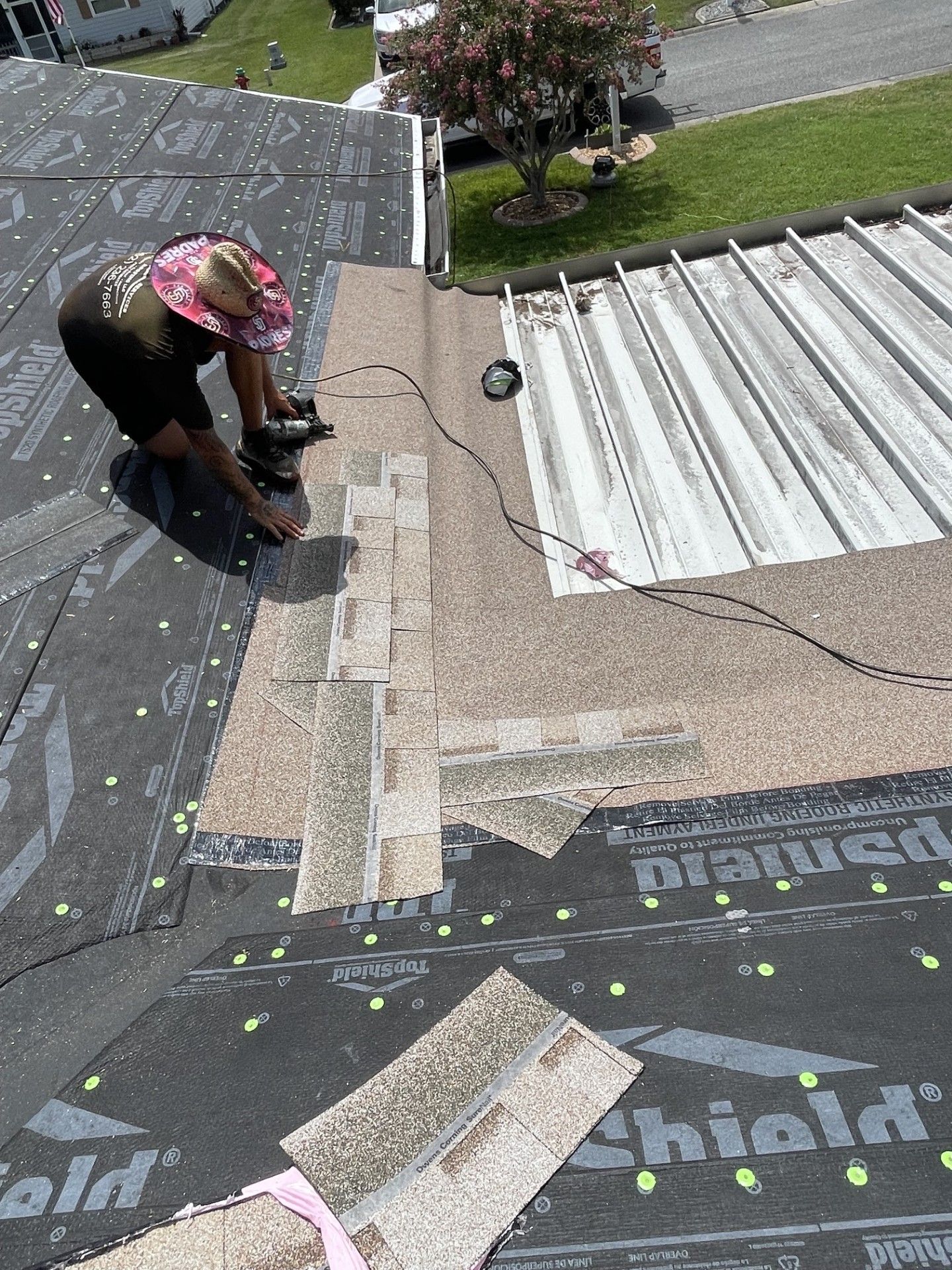 Worker on a rooftop, inspecting or repairing shingles near a white corrugated roof.