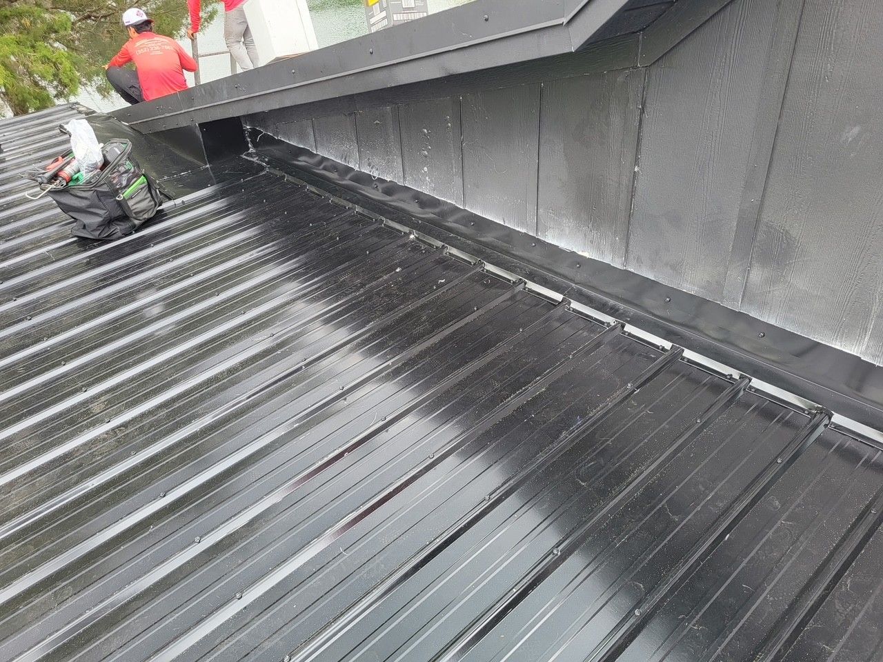 Workers installing a black metal roof on a sloped rooftop, with a raised wall along the side.
