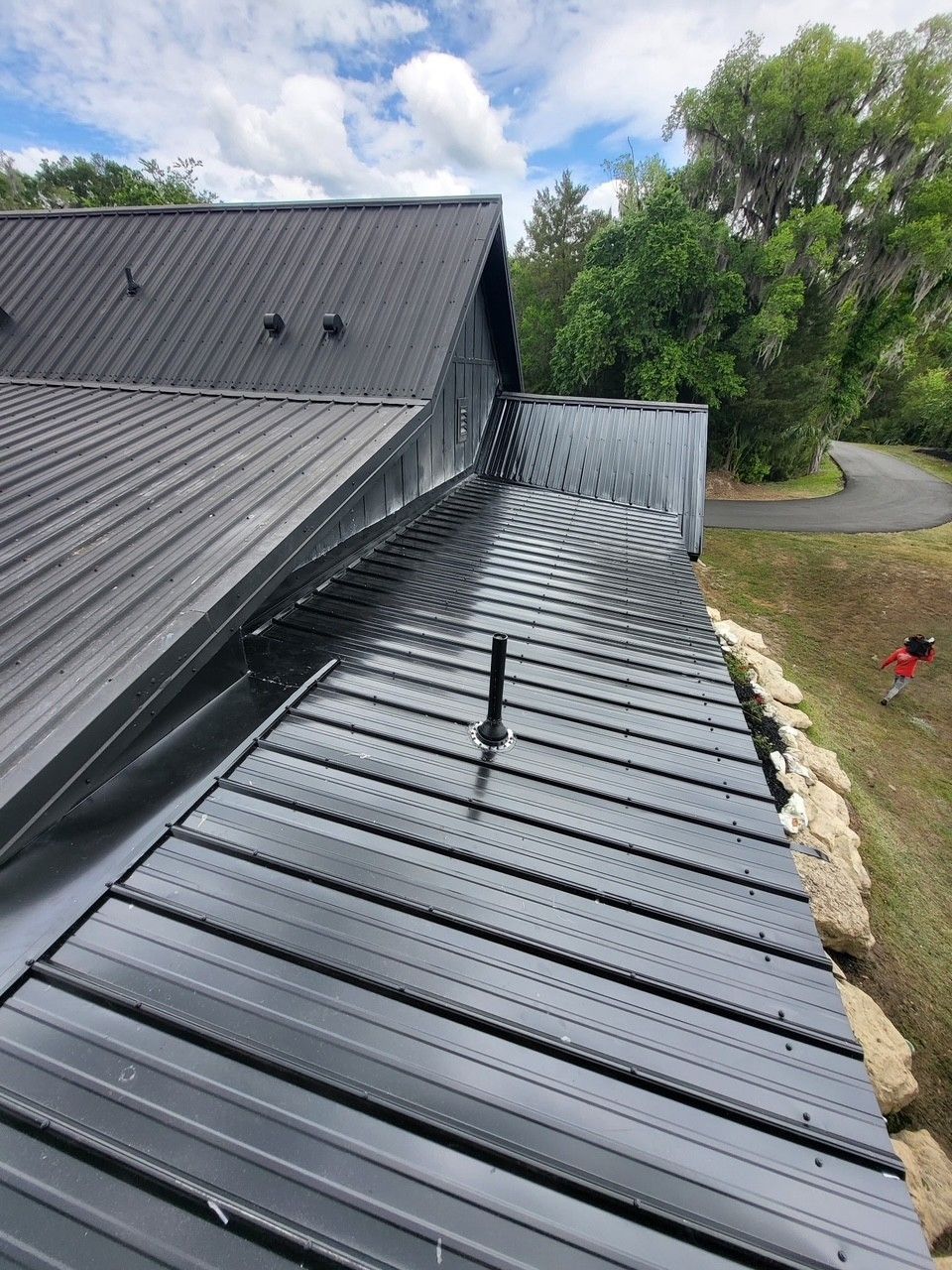 Black metal roof with standing seams beside a house and driveway under a cloudy sky