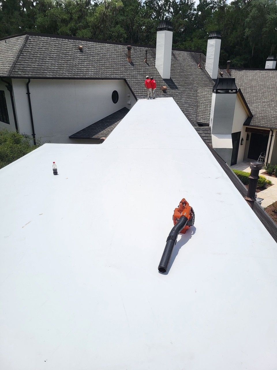 Roofer on a white roof near chimneys, with a chainsaw in the foreground and trees and houses behind.