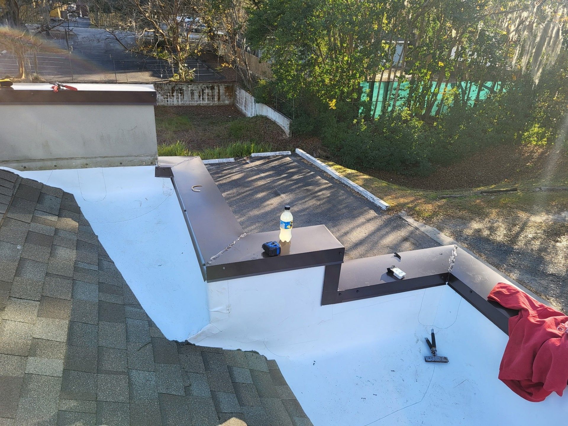 Rooftop under repair with white waterproof coating, shingles, tools, and a red tarp near trees.