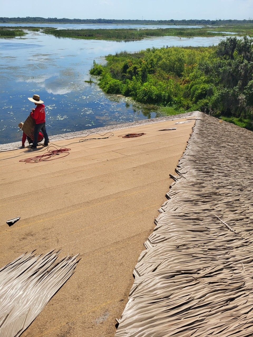 Person in a red shirt and white hat standing on a sandy shore beside a calm lake and green reeds.
