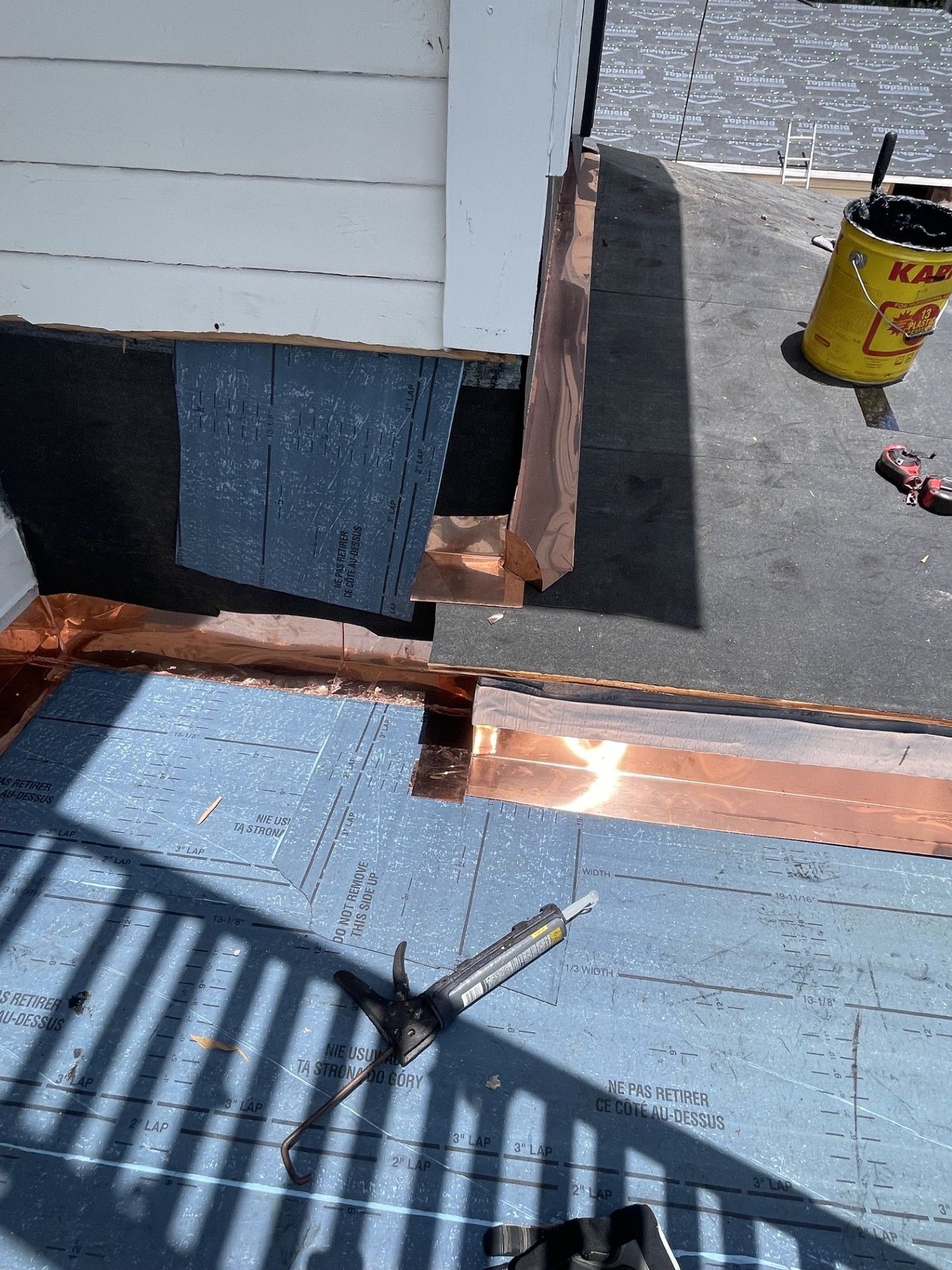Roof repair with blue shingles, exposed flashing, and a yellow bucket on a rooftop.