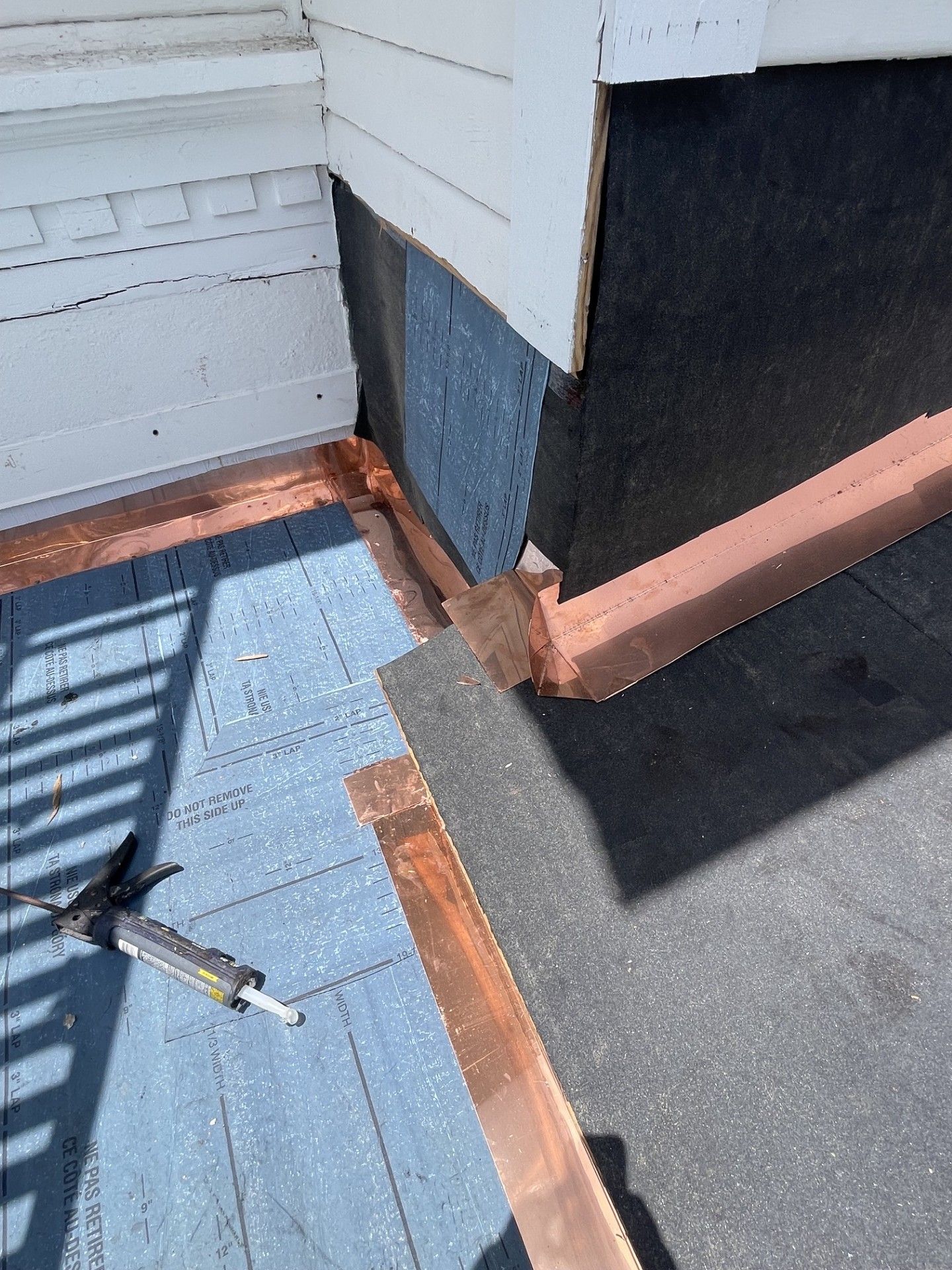 Corner of a roof with blue waterproofing, copper flashing, white siding, and tools on the surface.