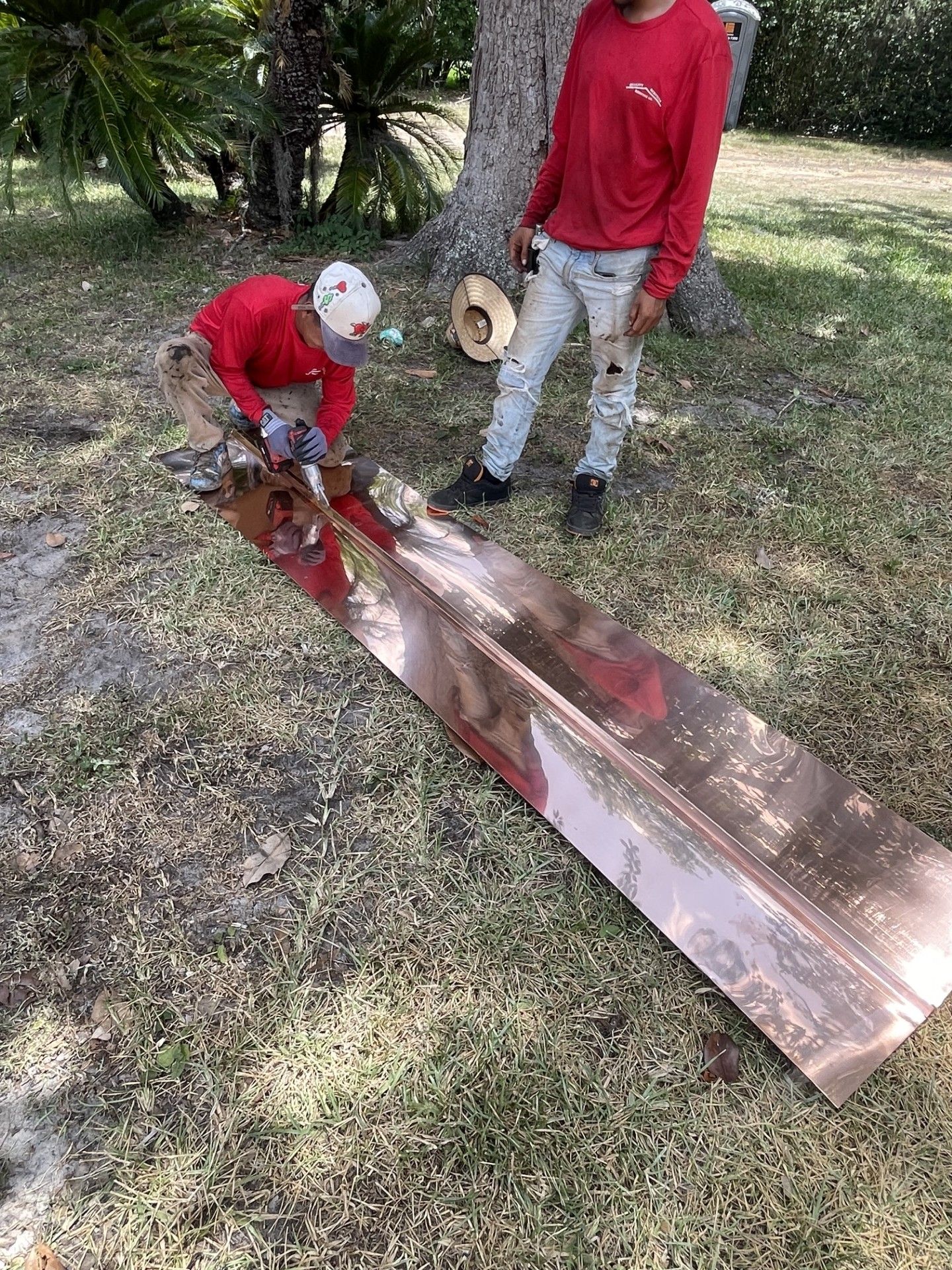 Two people in red shirts cutting a long wooden plank outdoors beside a tree.