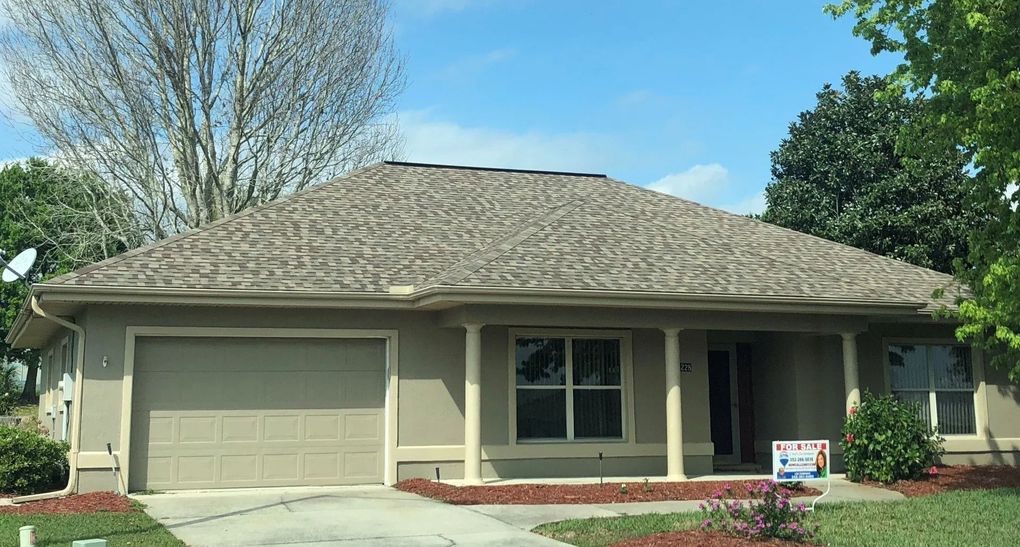 Single-story beige suburban house with a two-car garage, front porch, and a small lawn under a blue sky