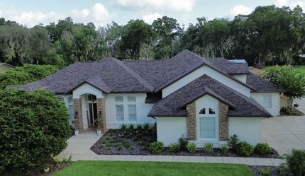 Large white suburban house with a dark roof, arched entry, and lush green trees around it.