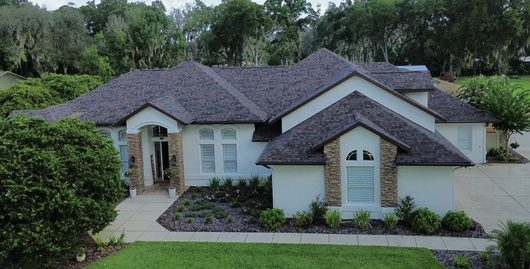 Large white suburban house with a dark roof, arched entry, and lush green trees around it.