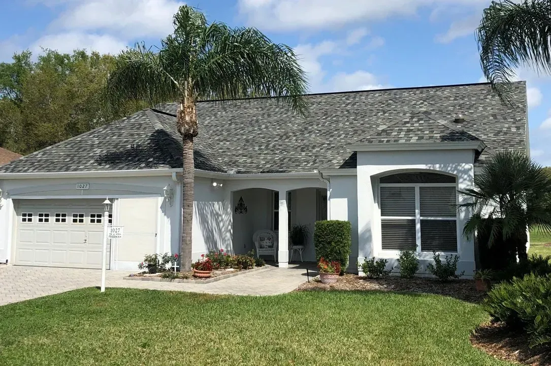 Single-story white house with gray roof, garage, palm tree, and green lawn under a blue sky