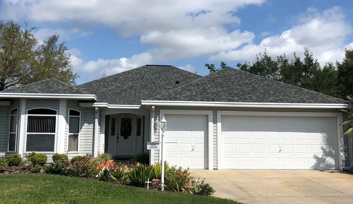 Single-story white house with a gray roof, front porch, flower beds, and a two-car garage.