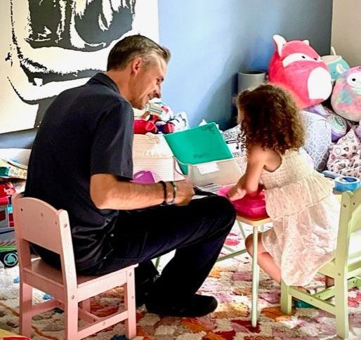 Man and child seated at small tables, looking at papers. Colorful playroom background.