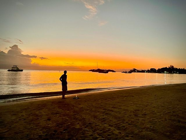 Silhouette of a person on a beach watching a fiery orange sunset over the calm ocean; a boat is visible.