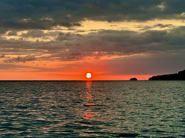 Sunset over calm ocean, orange and red hues reflecting in the water, dark clouds above.