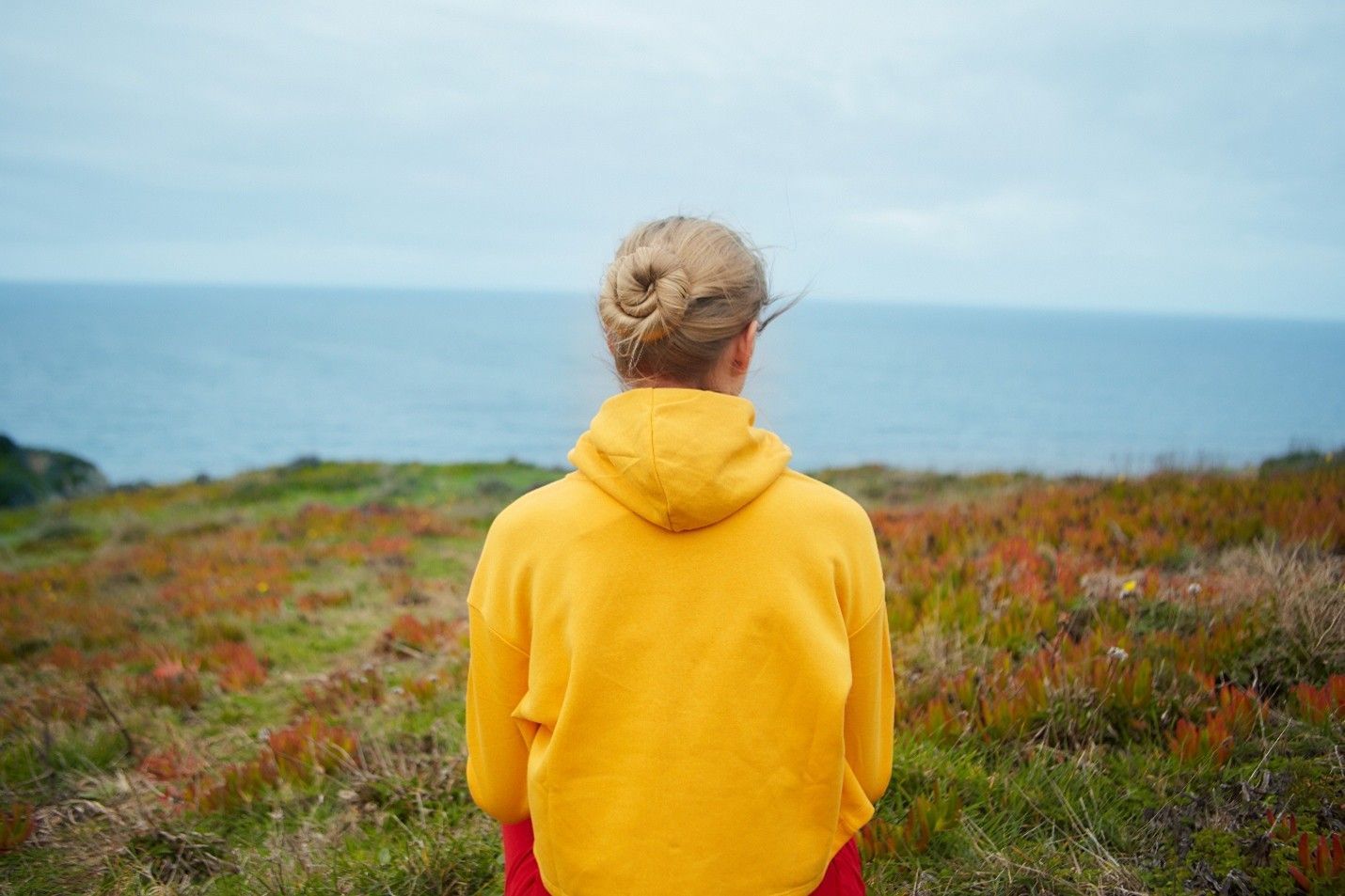 Person in a yellow hoodie looking at the ocean from a grassy cliff.