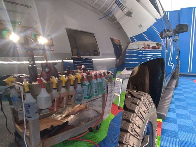 A rack of spray bottles sits in a garage next to a blue vehicle with a tire in the foreground.