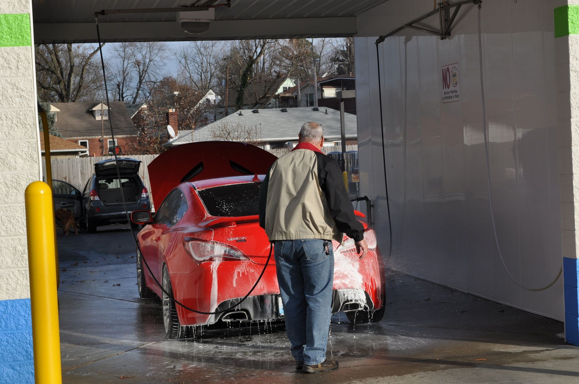 A person in a beige vest and jeans sprays a red car with an open hood at an outdoor car wash.