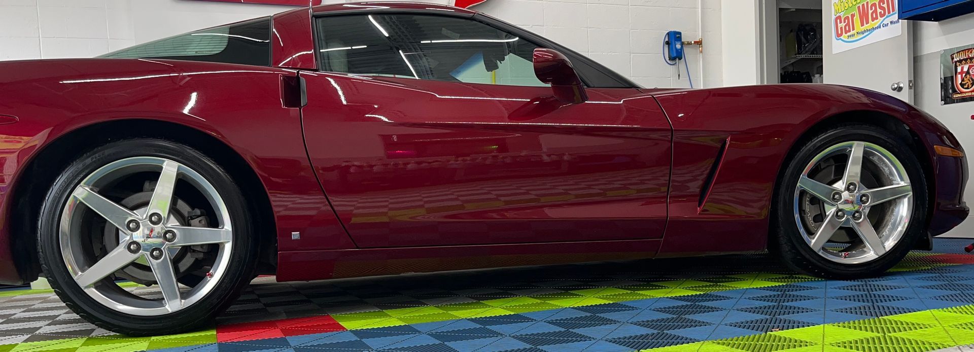 A side profile of a maroon C6 Chevrolet Corvette parked on a colorful, interlocking floor mat in a garage.