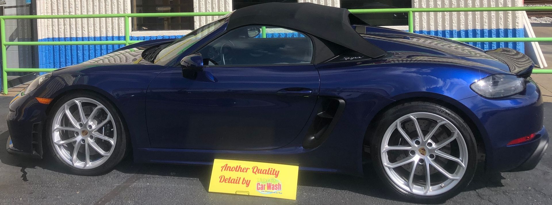 A blue Porsche convertible parked outdoors, with a small yellow sign placed on the pavement in front of the car.