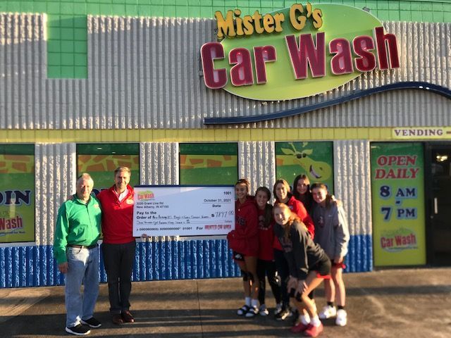 Two people stand next to a group of five holding a large check outside Mister G’s Car Wash under a sign reading Open Daily.