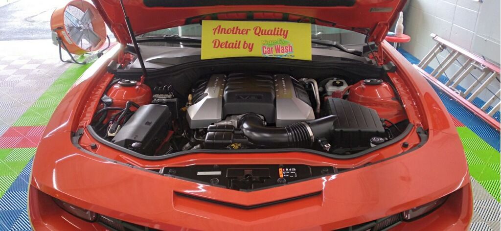 A bright orange Chevrolet Camaro with its hood open, revealing the engine bay in a garage with a colorful tiled floor.