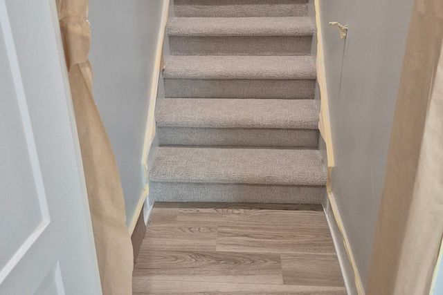 Narrow carpeted staircase with gray steps and wood flooring at the bottom, framed by protective covering on both sides