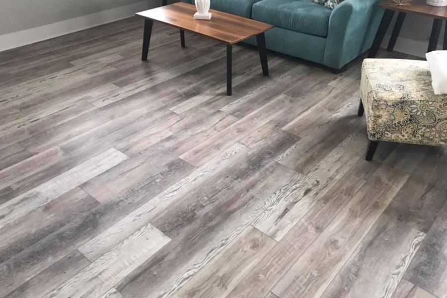 Modern living room with gray wood-look flooring, teal sofa, wooden coffee table, and floral ottoman.