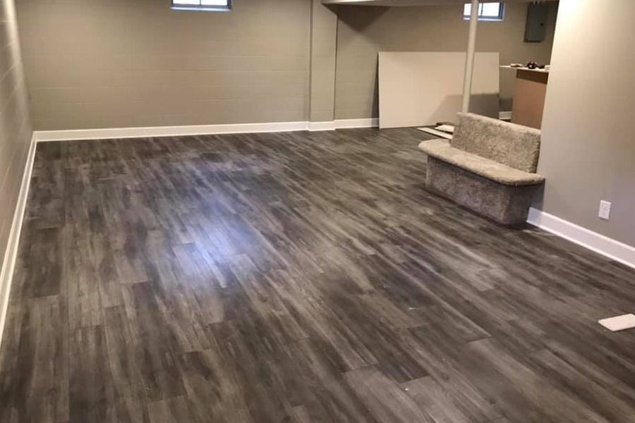 Empty basement room with gray wood flooring, beige walls, and a small chair near a doorway.
