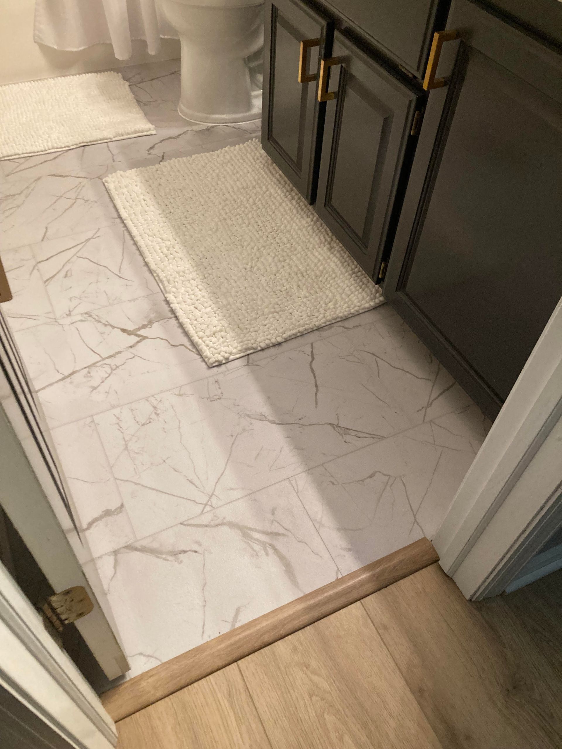 Bathroom doorway with a white marble tile floor, two bath mats, and a wood threshold beside dark cabinets.