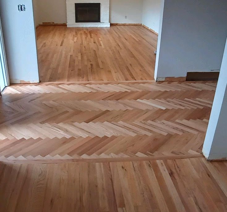 Empty room with hardwood floors and a herringbone wood inlay by a fireplace.