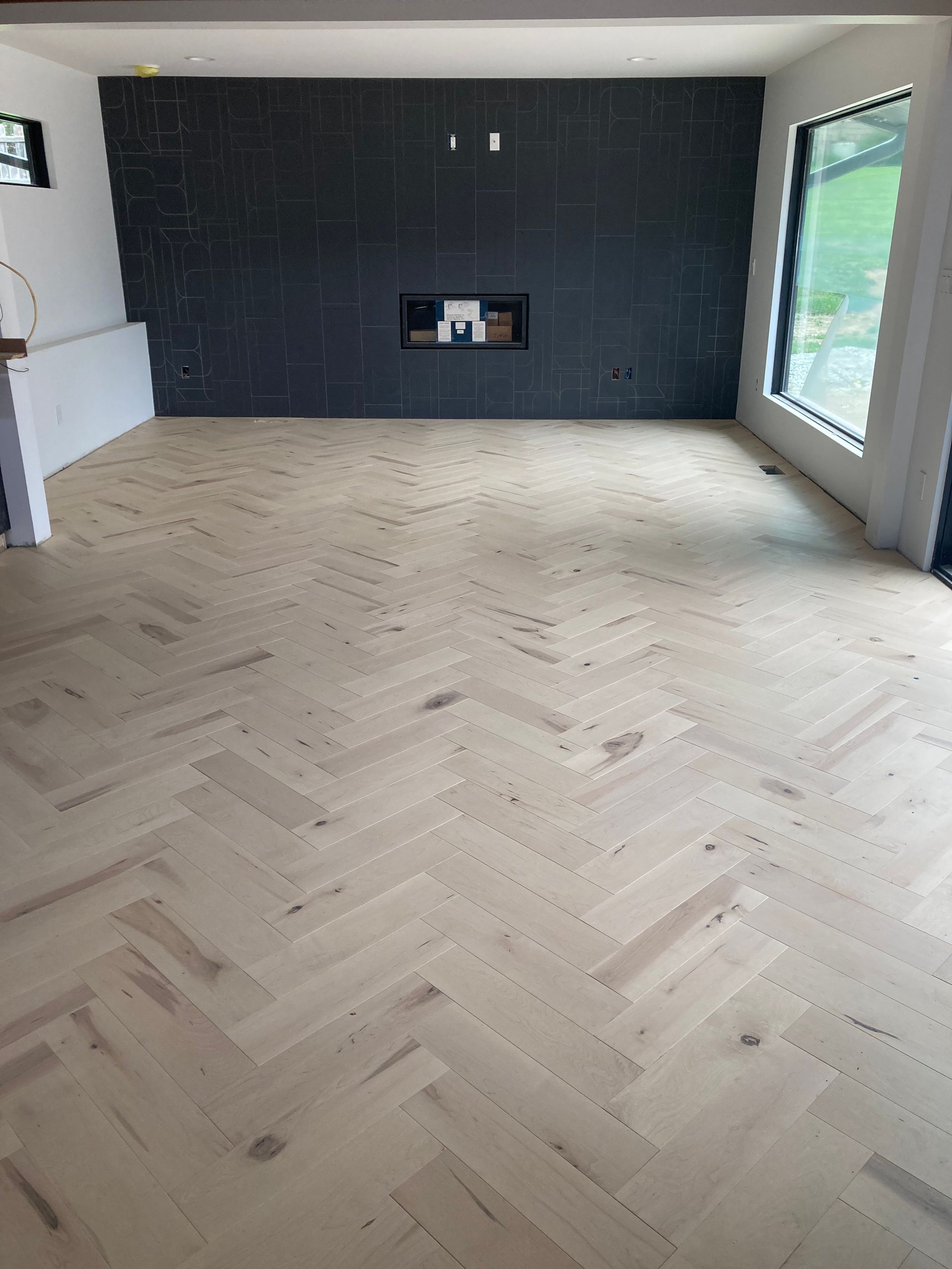 Empty modern room with light herringbone wood flooring and a dark fireplace wall.