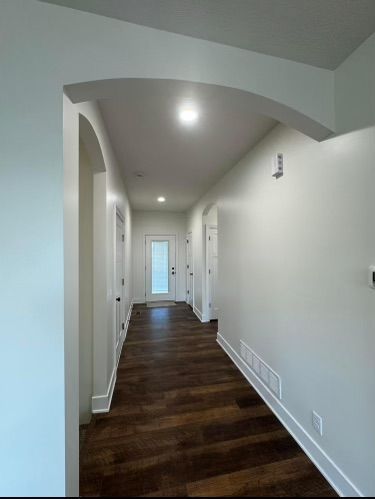 Narrow white hallway with dark wood floors and arched ceiling, lit by recessed lights, leading to a door.