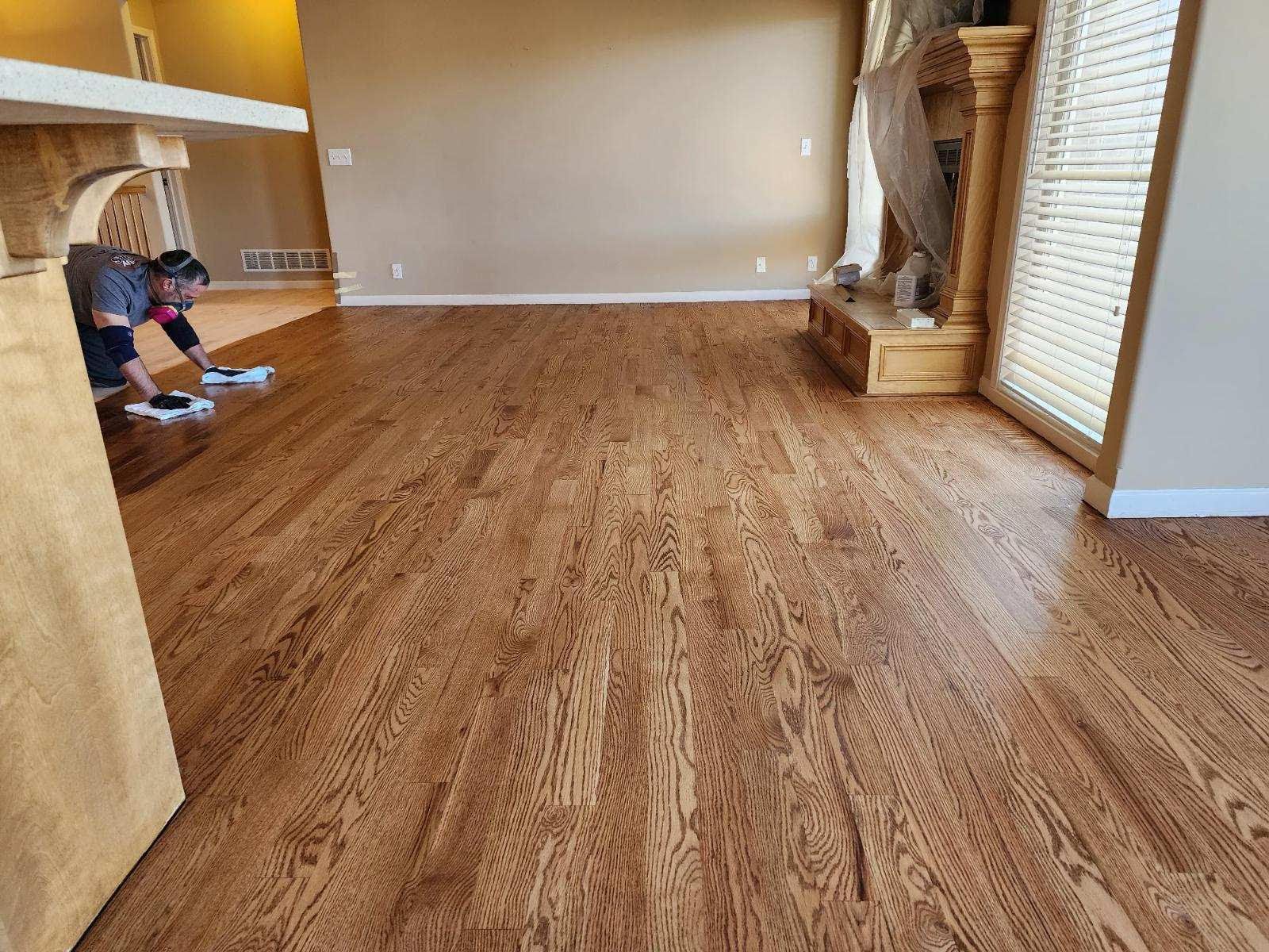 Person working on hardwood flooring in a beige living room with stone fireplace and large windows