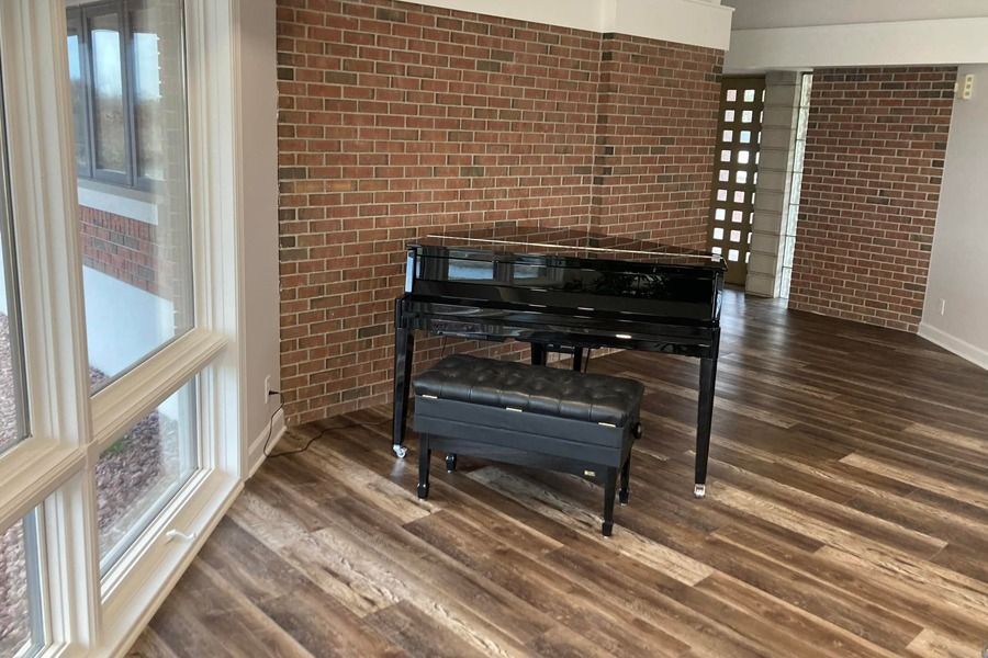 Black upright piano in a brick-walled room with wood floors and large windows