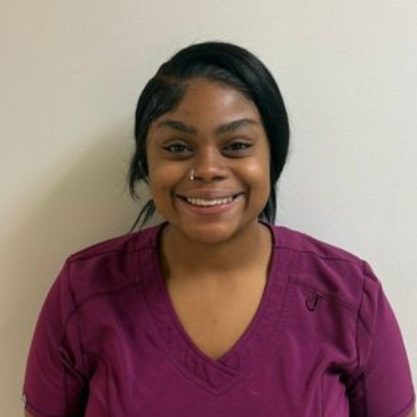 Woman in black scrubs, smiling, with braided hair, standing in front of a white wall.