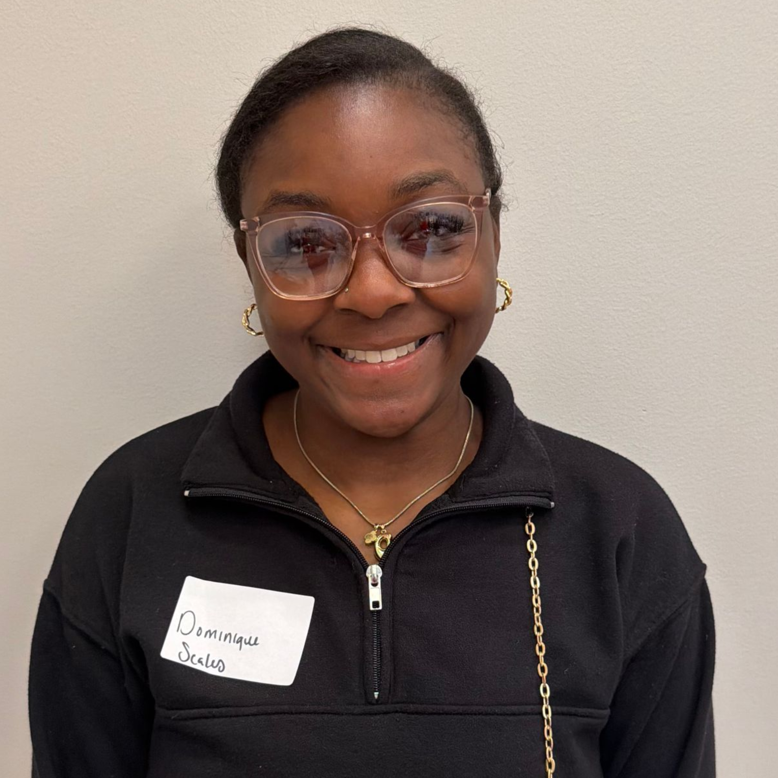 Woman with glasses and a zip-up sweater smiles in front of a white wall.