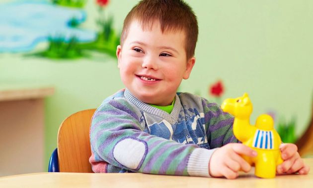 Boy with Down syndrome smiling, holding a yellow toy camel at a table.