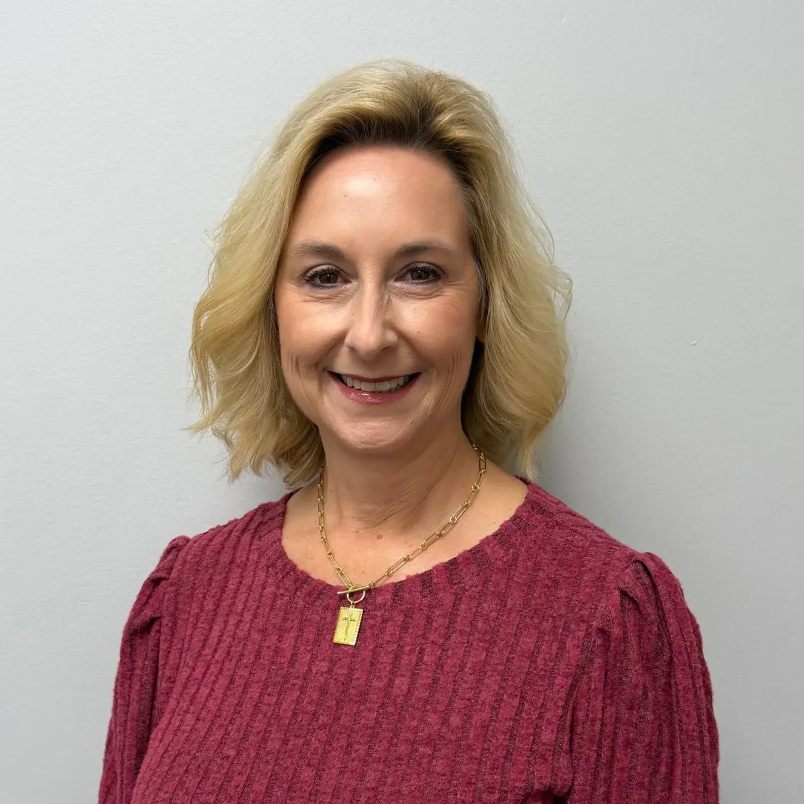 Woman with blonde hair wearing a maroon sweater and gold necklace smiles in front of a gray wall.