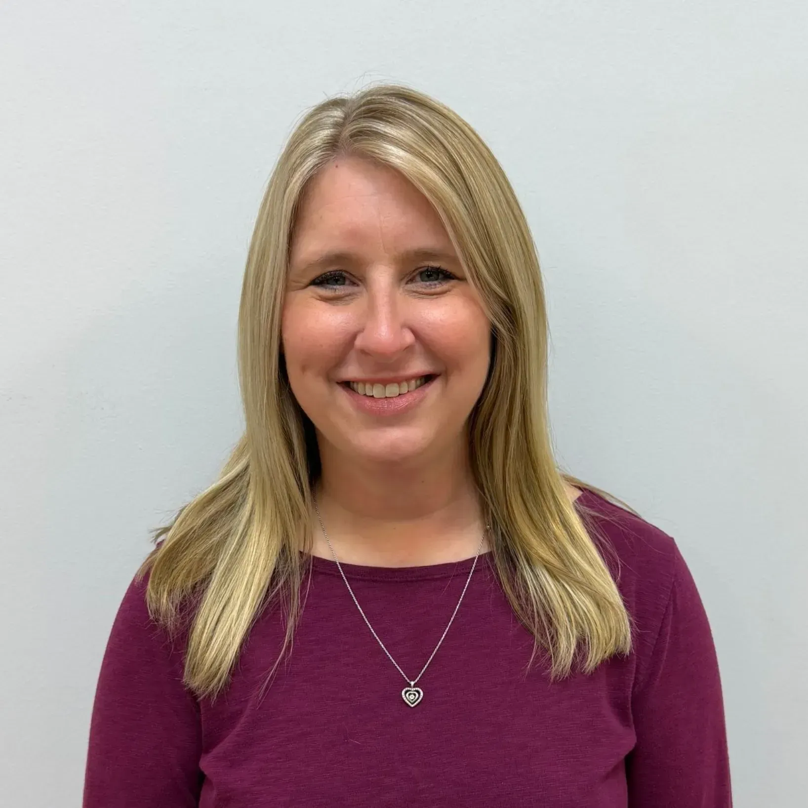 Woman with blonde hair wearing a maroon top and silver heart pendant, smiling against a white background.