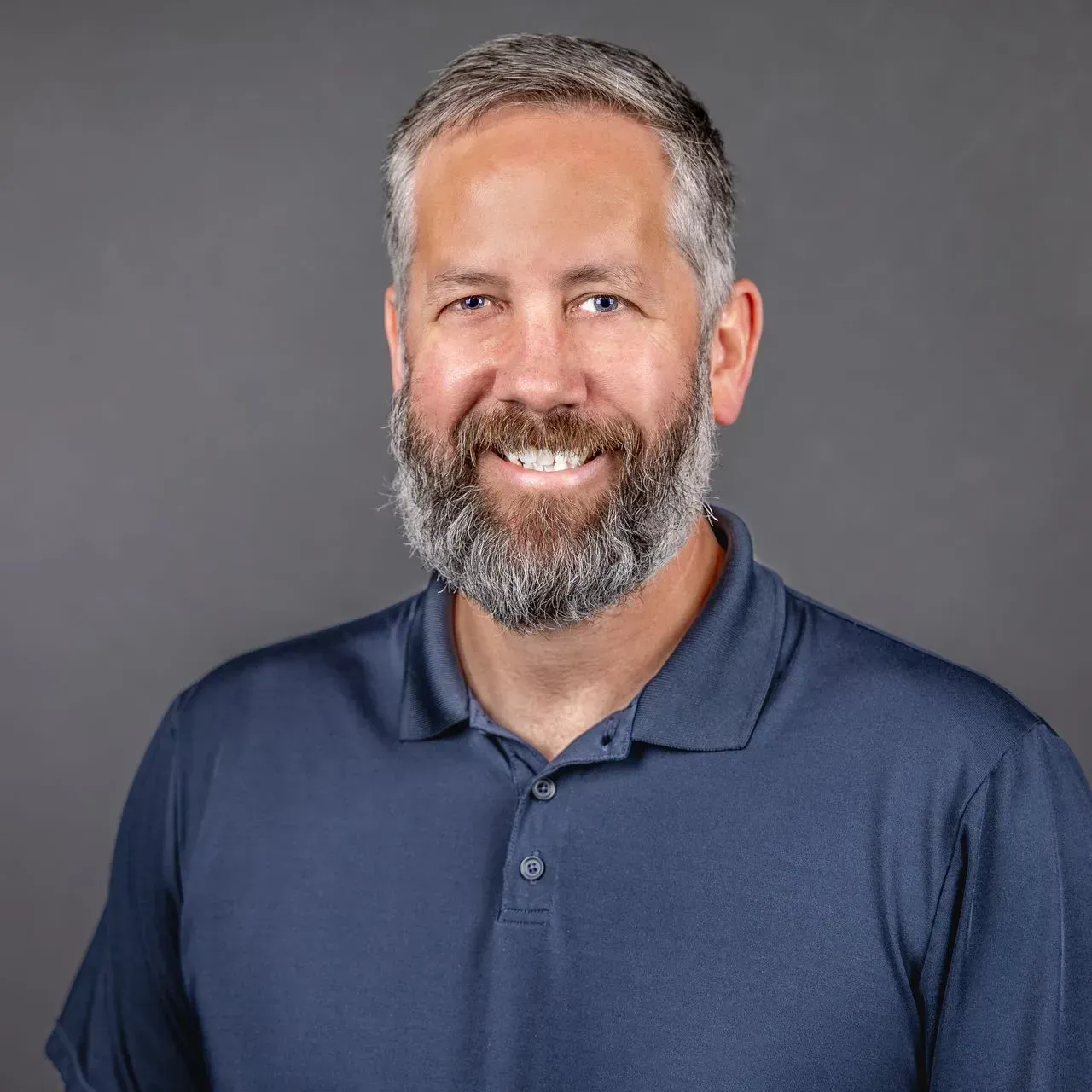 Man with a salt and pepper beard smiles, wearing a navy blue polo shirt against a gray backdrop.